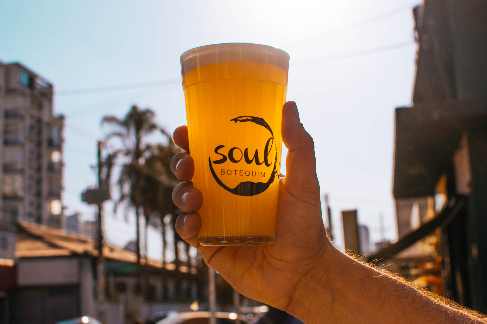 Mão erguendo um copo de chope do Soul Botequim contra o sol do fim de tarde
